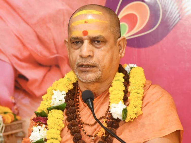 माइक्रोफ़ोन में बोलते हुए स्वामी सदानन्द सरस्वती का निकट से लिया गया चित्र; माथे पर पीला और लाल तिलक, तथा गेंदे, चमेली और रुद्राक्ष की मालाएँ