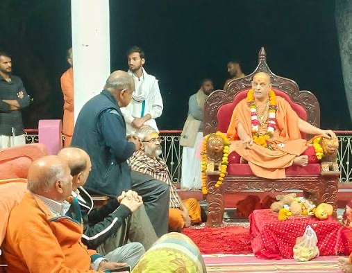 The Shankaracharya seated on a carved throne with lion figures at night while devotees sit before him and one addresses him