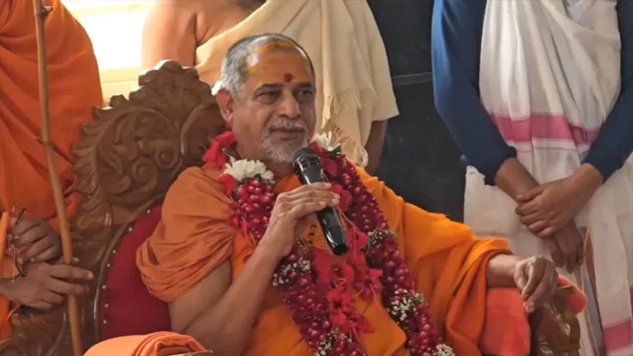 Swami Sadanand Saraswati speaking into a handheld microphone from a carved wooden chair, garlanded with red roses