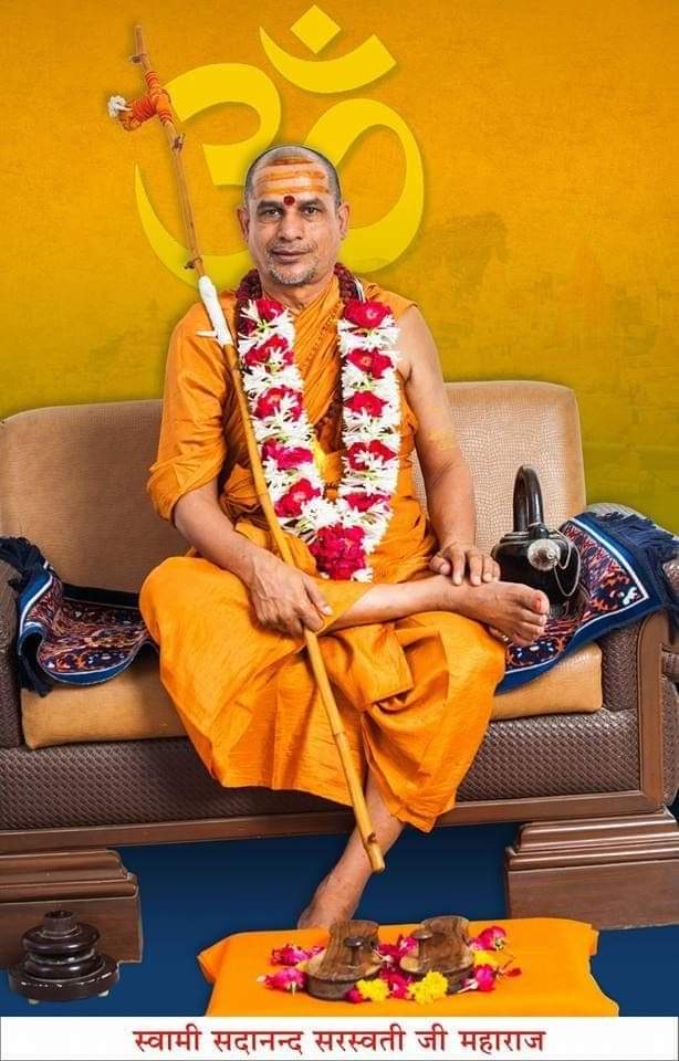 Swami Sadanand Saraswati seated on a sofa in saffron robes, holding a danda, garlanded with red and white flowers, before a saffron backdrop with Om, with his name in Devanagari at the foot