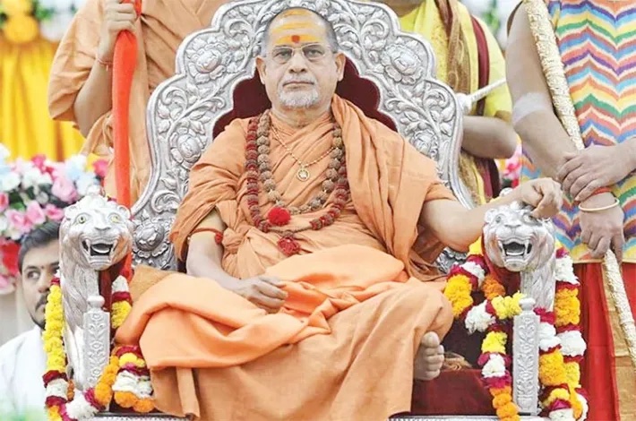 Swami Sadanand Saraswati in spectacles and rudraksha seated on an ornate silver throne with lion armrests and marigold garlands