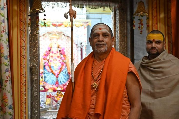 Swami Sadanand Saraswati standing in a saffron shawl with a danda before a decorated shrine of the Goddess