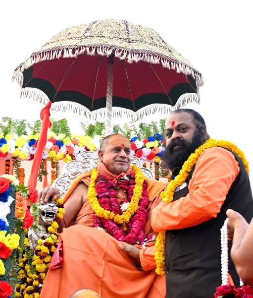 The Shankaracharya smiling on a silver throne under a maroon and gold parasol with a garlanded devotee beside him