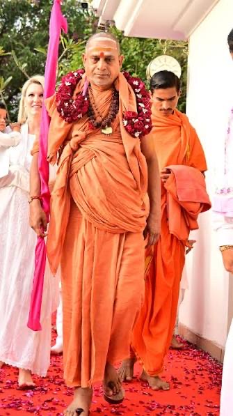 Swami Sadanand Saraswati walking barefoot on rose petals in a saffron robe with a garland of red roses, followed by a young monk