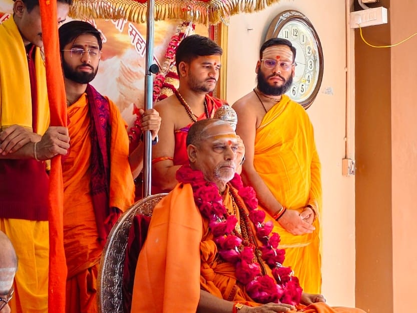 Swami Sadanand Saraswati seated with a garland of red roses while young disciples in saffron and yellow stand behind him