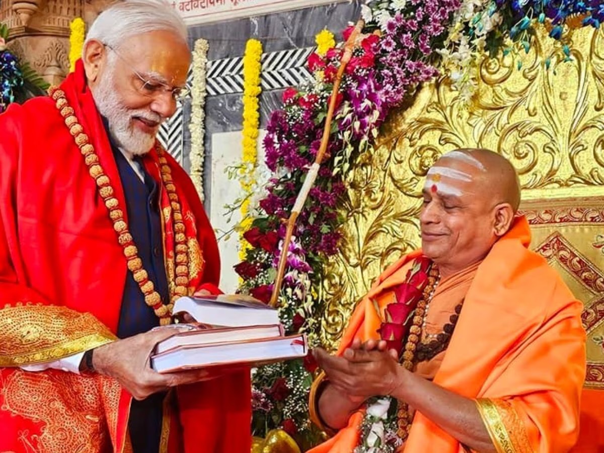 Prime Minister Narendra Modi, in a red stole and rudraksha mala, presents a set of books to the Shankaracharya, who smiles with hands raised, before a gold-leaf backdrop decked with orchids