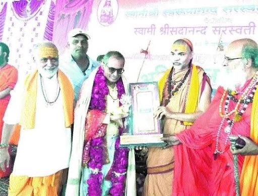 Newspaper photograph of a felicitation on a stage: sannyasis in saffron present a framed memento to a garlanded guest, below a banner reading Swami Shri Sadanand Saraswati in Devanagari