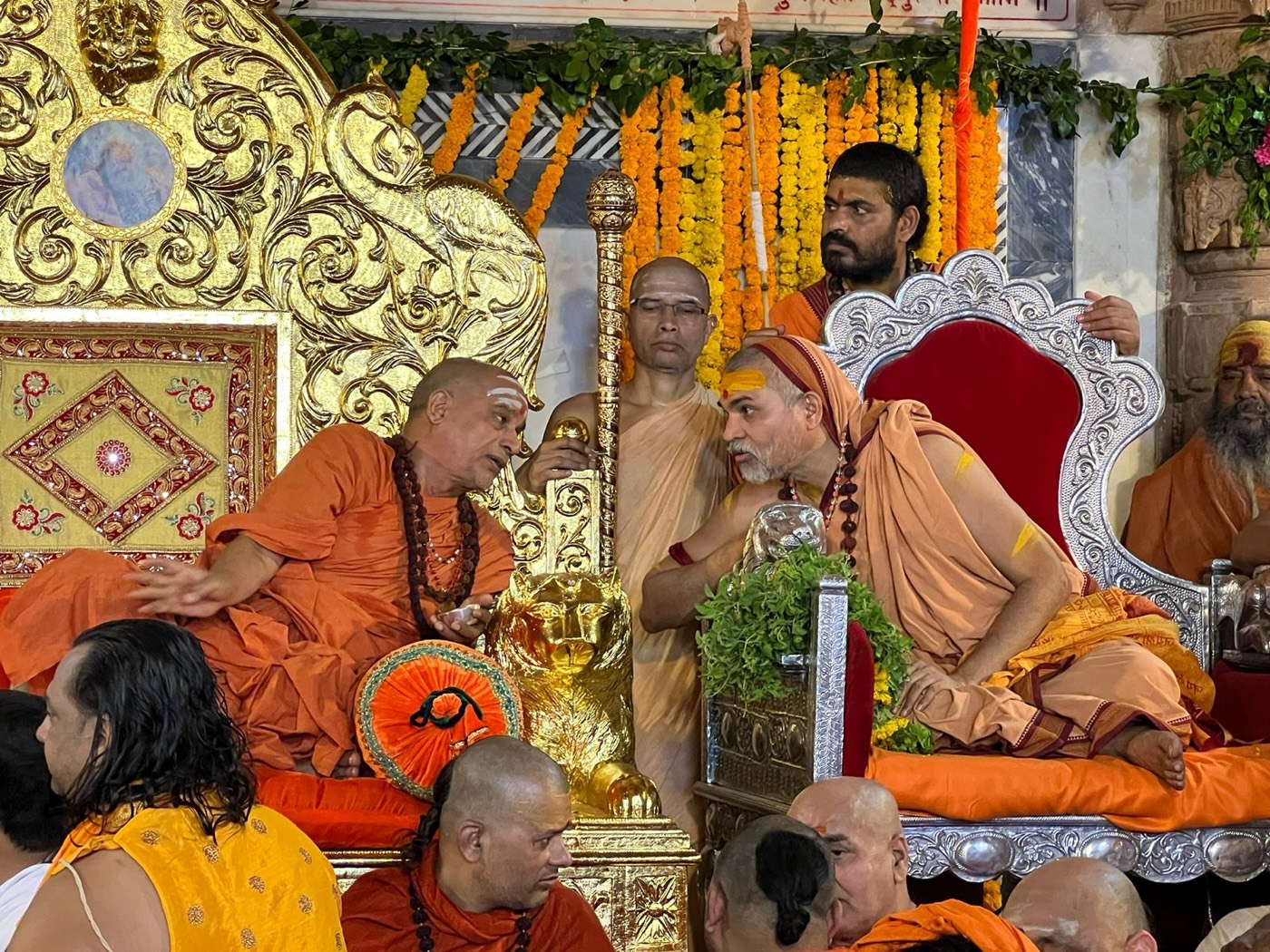 Two acharyas in saffron seated on ornate gold and silver thrones, leaning together in conversation, surrounded by attendants, marigold garlands and a golden lion-headed staff